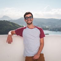 Young man in sunglasses leaning on a balcony with a scenic city view in the background.
